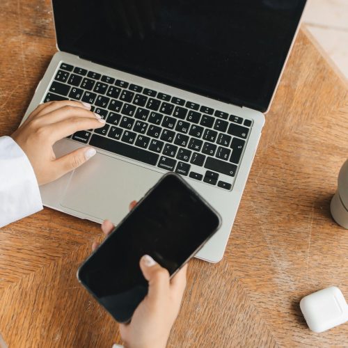 Person using a laptop and smartphone simultaneously at a wooden table.
